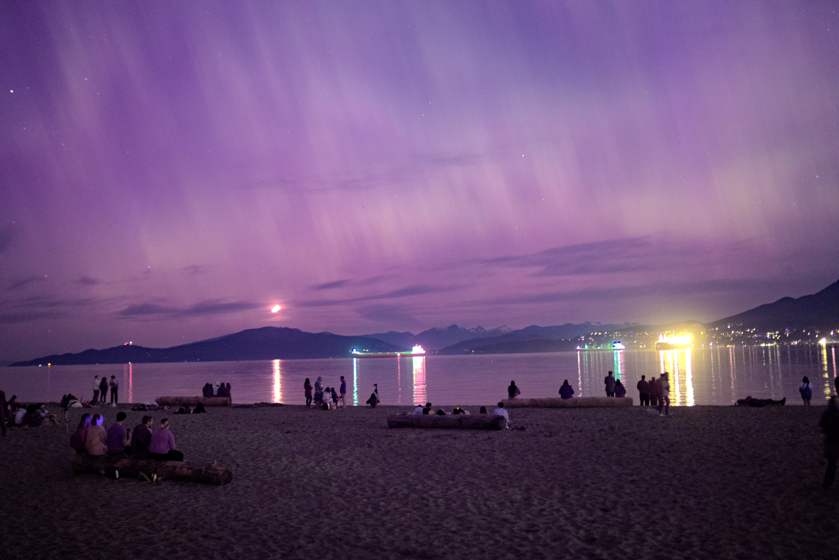 A nighttime beach scene under a vibrant purple sky. Silhouettes of people on the beach contrast with the lights reflecting on the water.