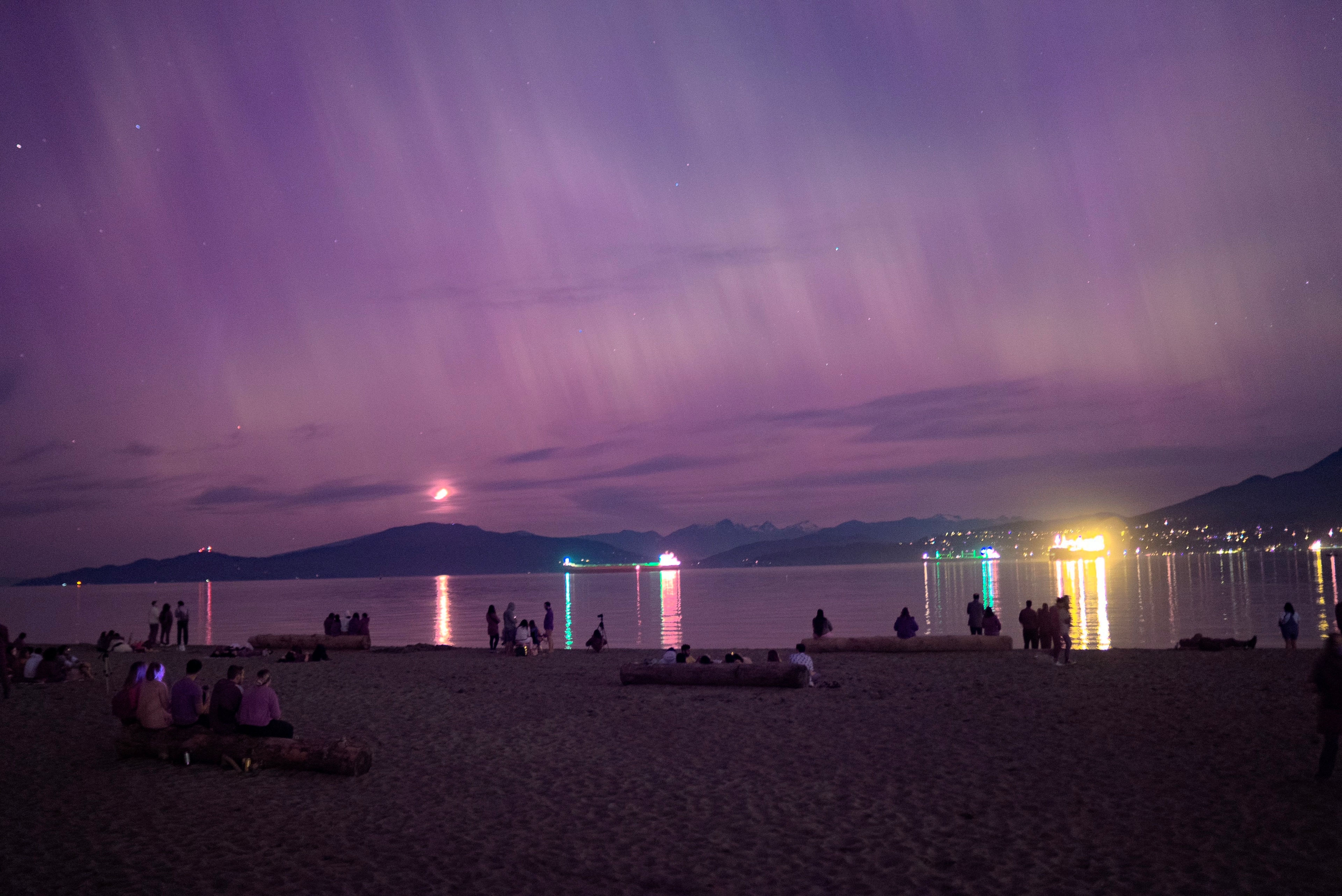 A nighttime beach scene under a purple sky, with people silhouetted against reflected light on the water.