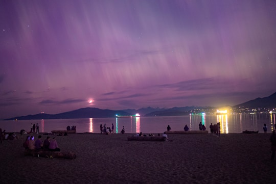 A nighttime beach scene under a purple sky, with people silhouetted against reflected light on the water.