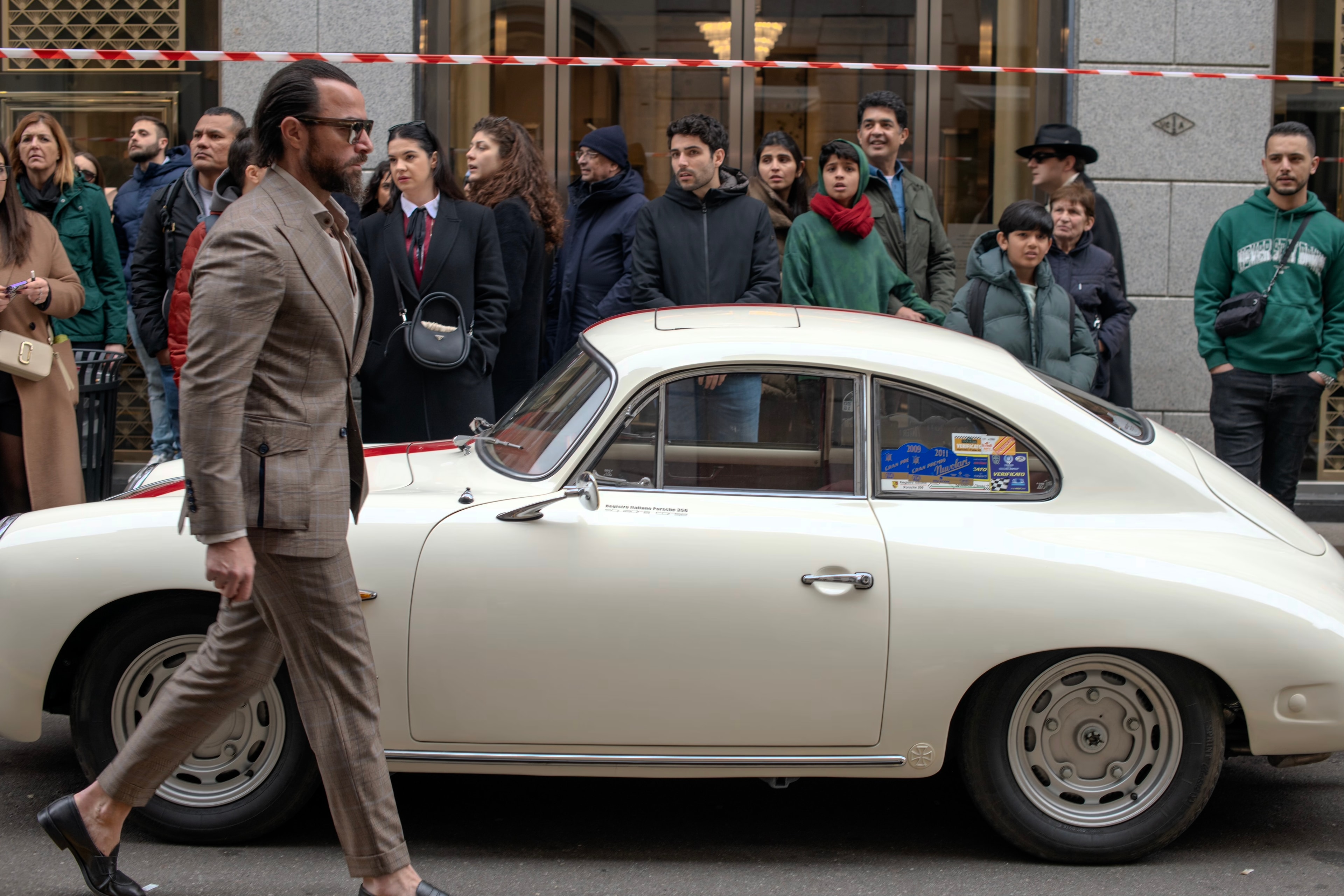 A well-dressed man walks past a vintage cream-colored Porsche. A crowd of people watch the scene.