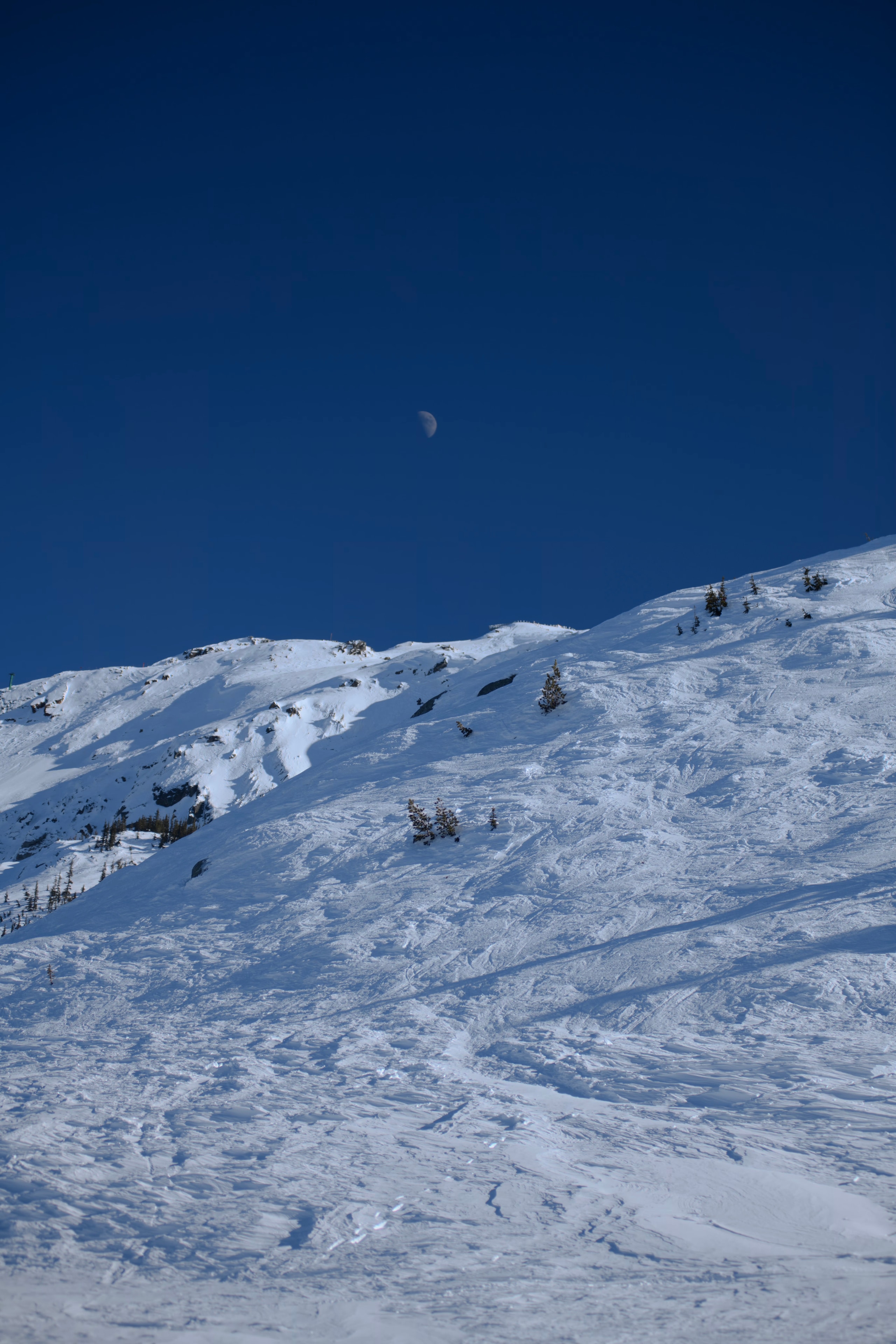 A snowy mountain landscape under a clear blue sky. A crescent moon is visible in the sky.