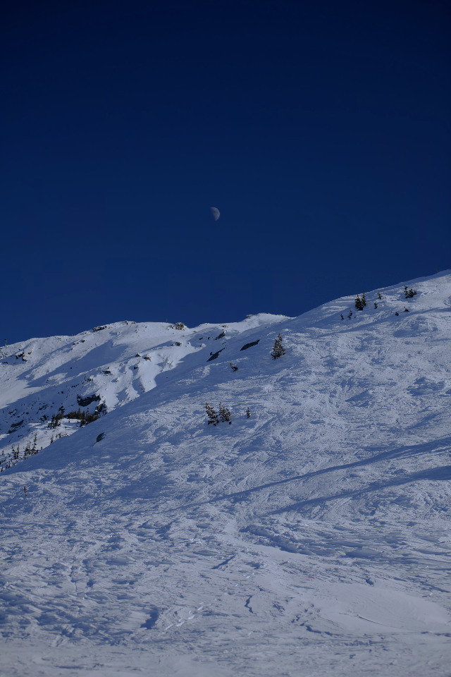 A snowy mountain landscape under a clear blue sky. A crescent moon is visible in the sky.