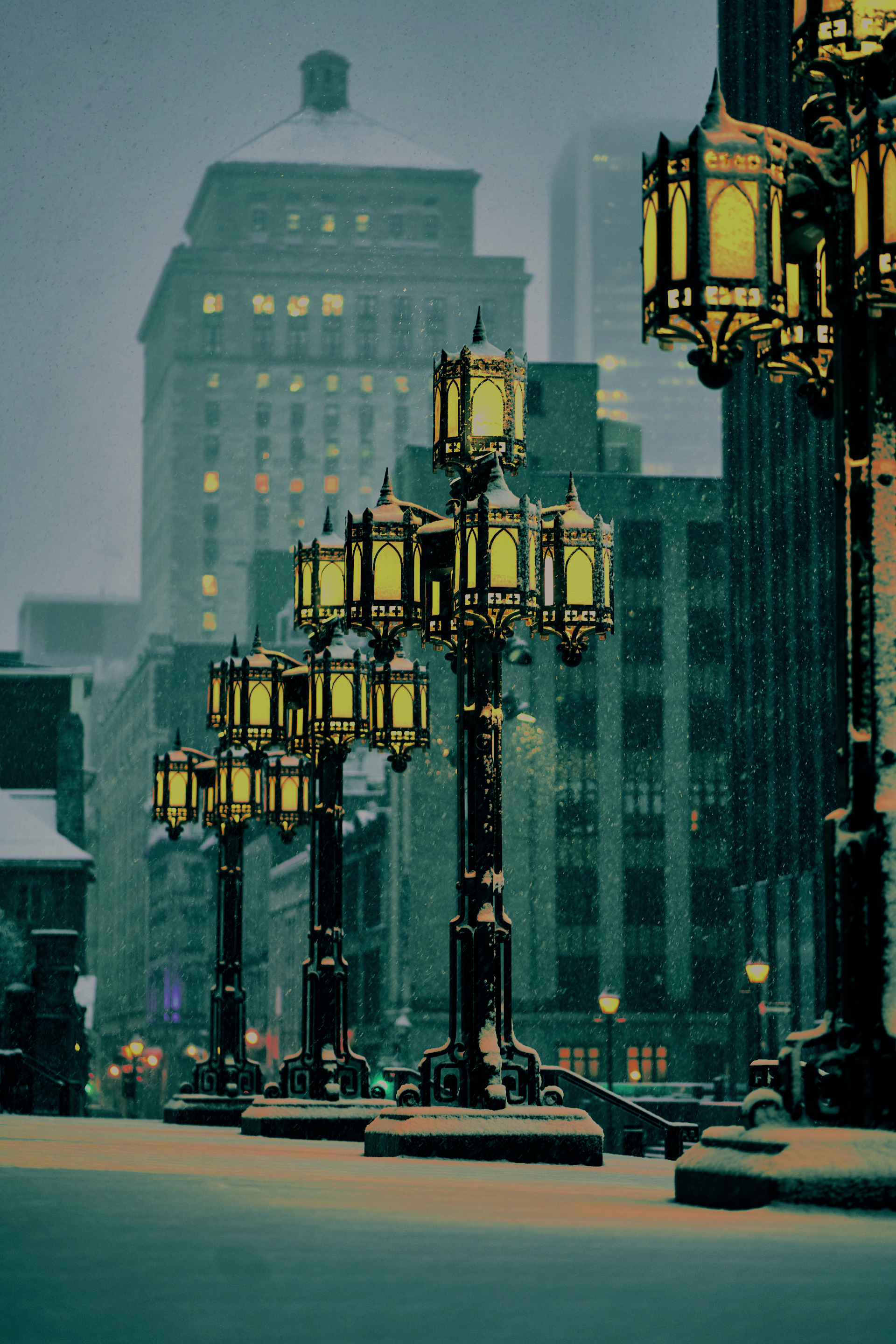 A nighttime winter scene with ornate streetlights illuminated against a backdrop of buildings. Snow falls gently, creating a serene and atmospheric ambiance.