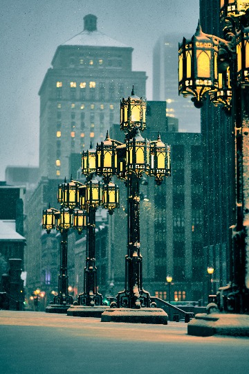 Snow falls through ornate streetlights at night, with buildings behind them.